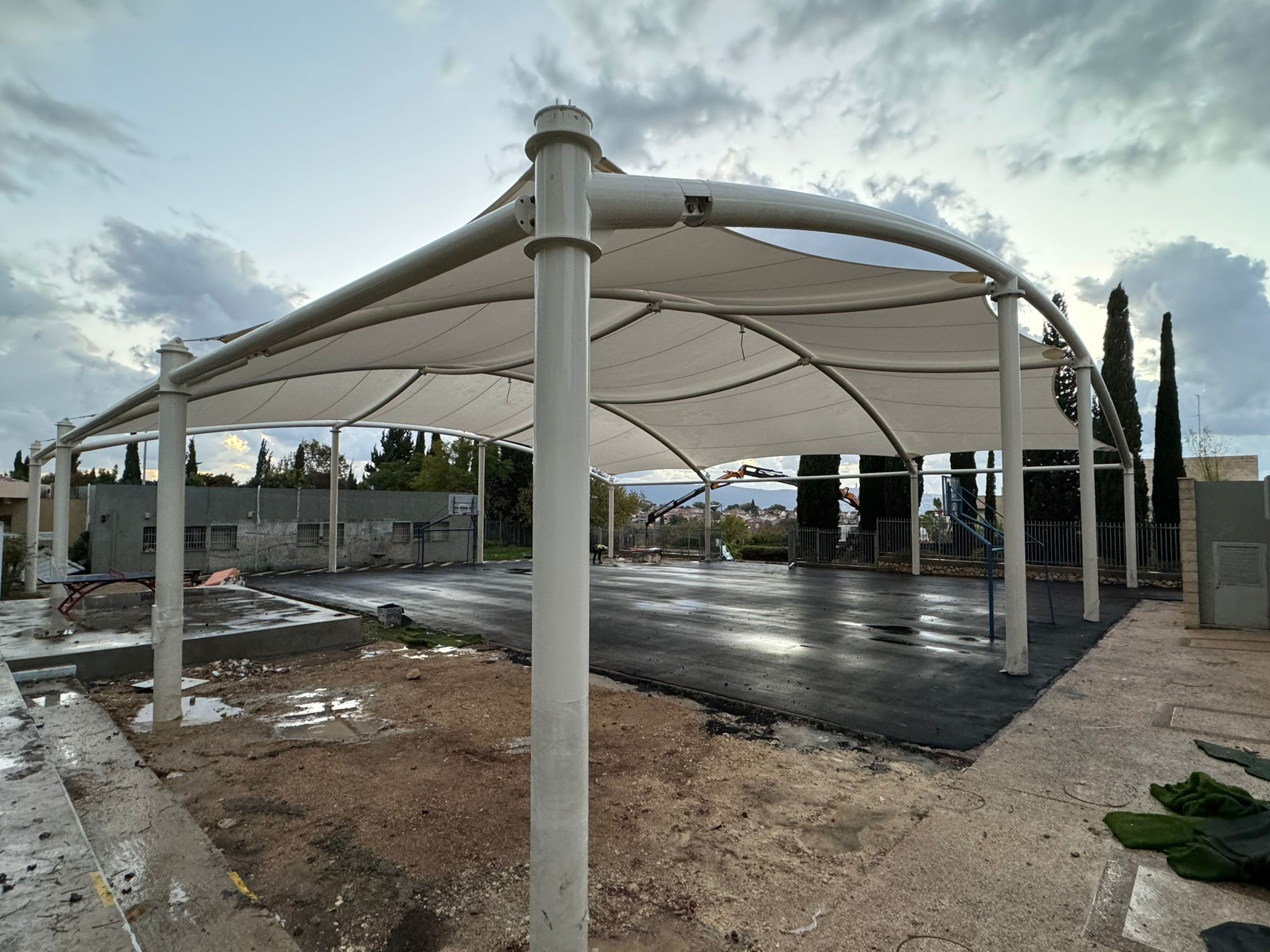 Canopy with beige stretched fabric roof on light-coloured metal supports over paved area under cloudy sky.