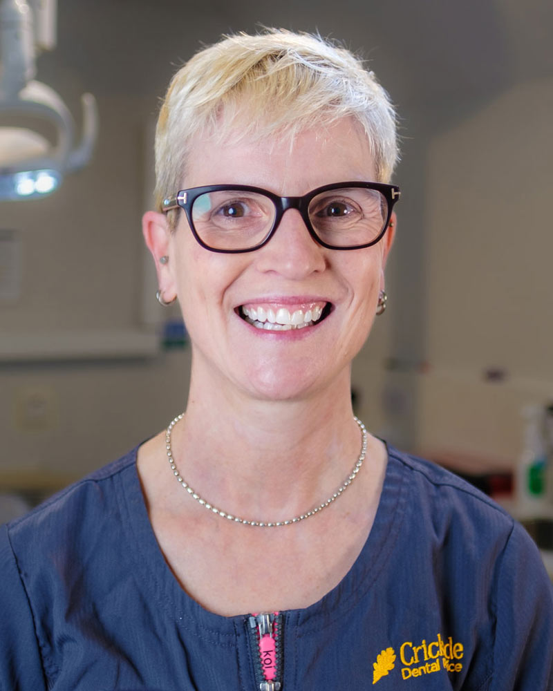 A portrait of Tracey, a Dental Nurse at Cricklade Dental Practice, smiling and wearing dark-framed glasses and a dark grey scrub top with a pink zipper pull. She is wearing a name badge that reads "Tracey" and the top features yellow embroidered text reading "Cricklade Dental Practice" on the right. She is standing in a dental surgery with a dental light visible in the background.