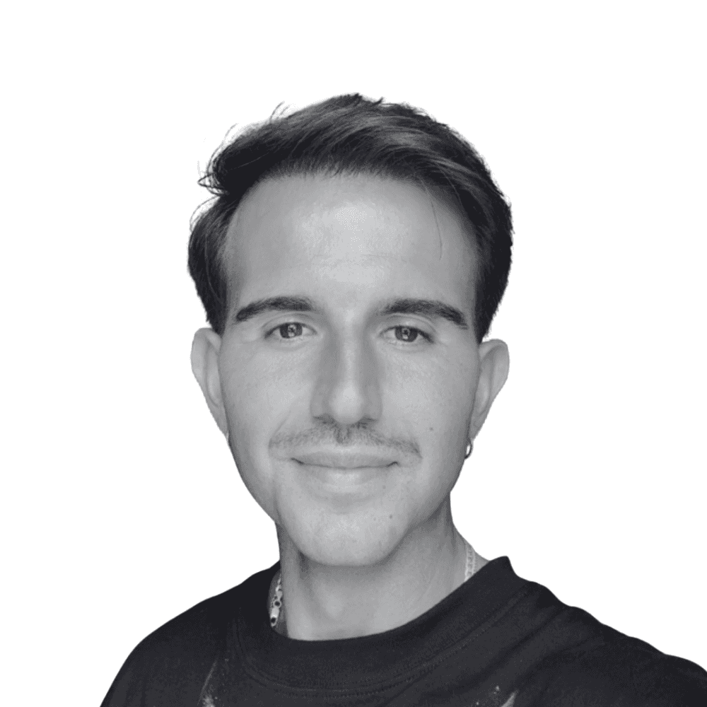 Asian man with glasses in a creative workspace, surrounded by colorful abstract art and stationery on a cluttered desk.