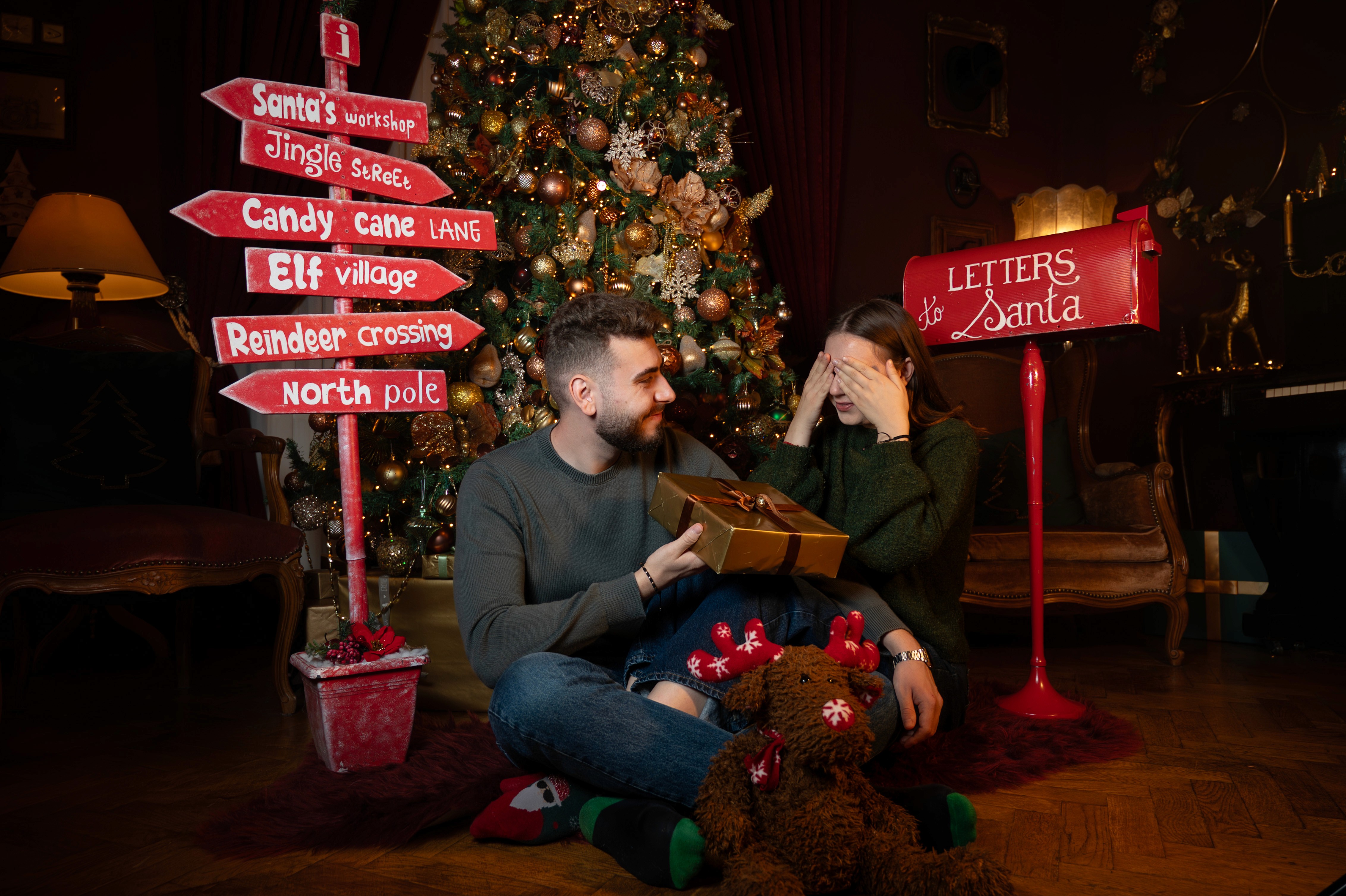 Ședință foto de Crăciun cu cuplu într-un decor festiv, indicatoare tematice și brad decorat