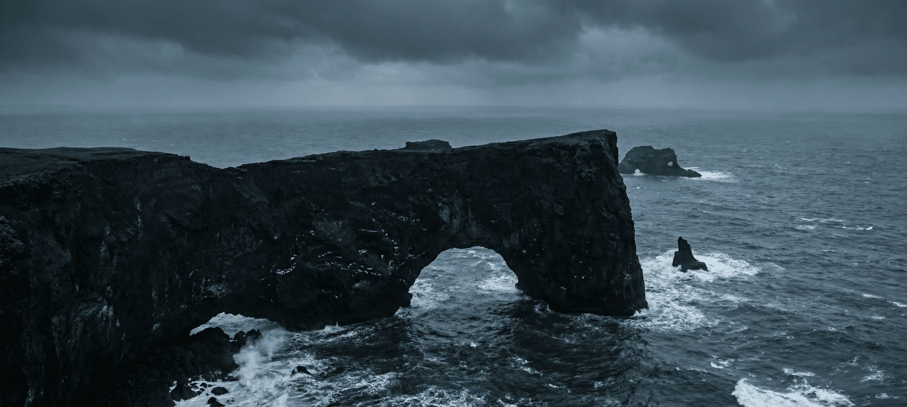 A large natural rock arch standing in the sea under a moody, cloudy sky.