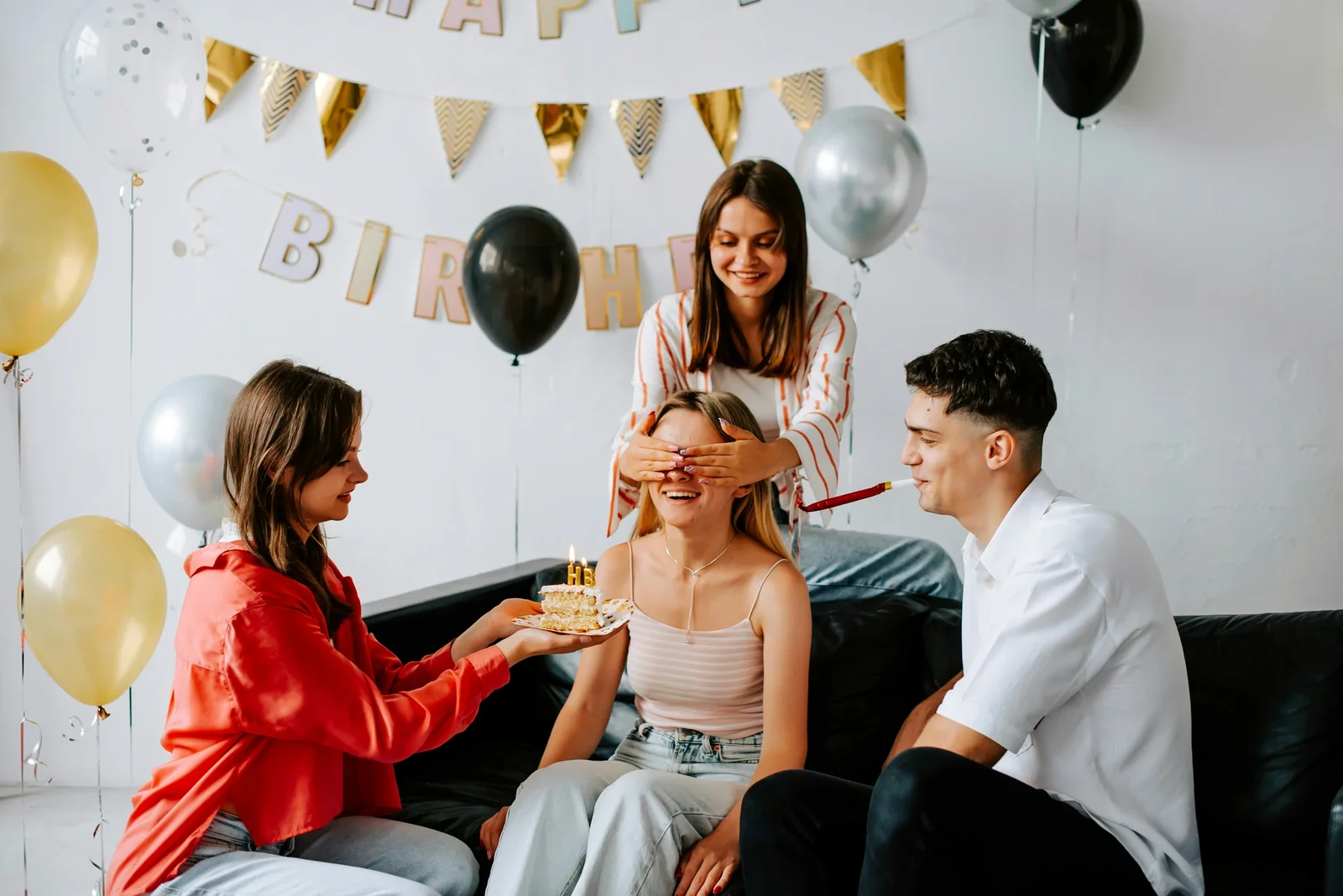 Friends surprise birthday girl with cake and party decorations featuring "HAPPY BIRTHDAY" banner and balloons.