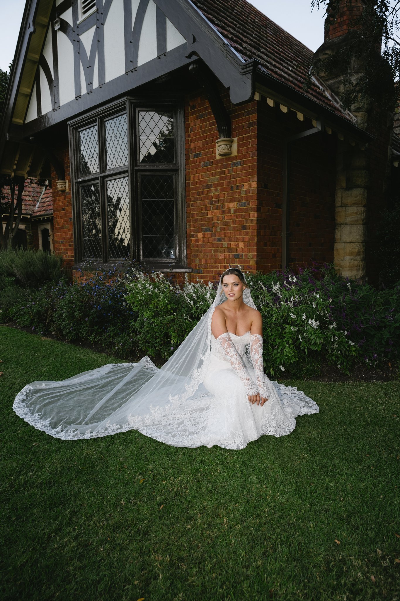 Bride seated on lawn with long lace veil spread out, in front of a Tudor-style brick cottage and garden