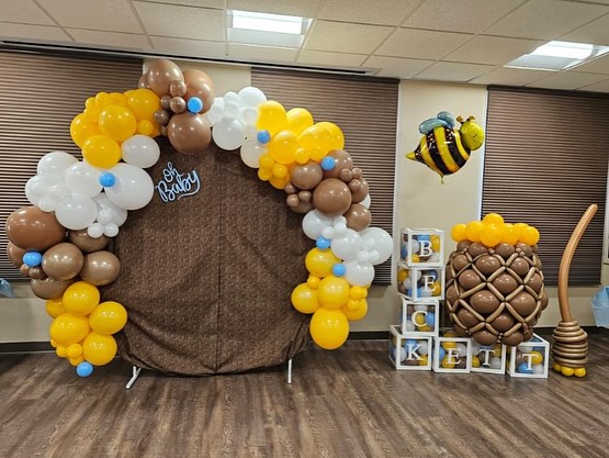This image shows a vibrant baby shower setup featuring a large balloon arch in shades of yellow, brown, and white, a bee-themed decoration, blocks spelling "Beckett," and a giant honey dipper, creating a cheerful and festive atmosphere.