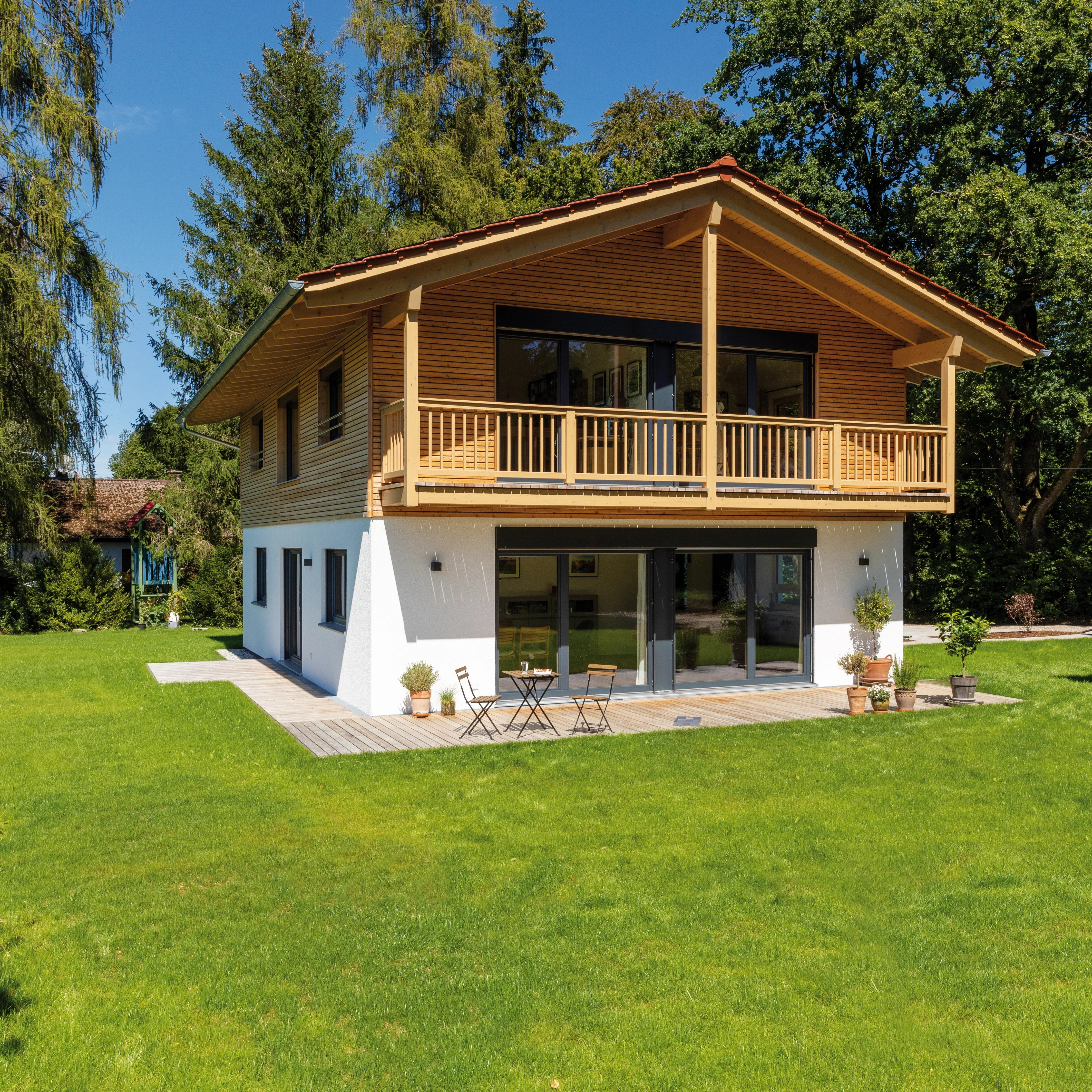 Modernes Holzhaus mit weißer Fassade, Balkon und großer Terrasse, umgeben von einer gepflegten Rasenfläche und Bäumen.
