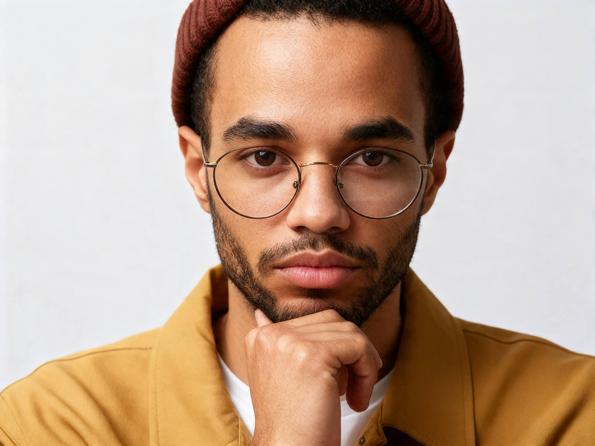 Smiling young designer wearing glasses and a burgundy beanie, dressed in a light t-shirt and caramel jacket, looking to the side against a neutral background.