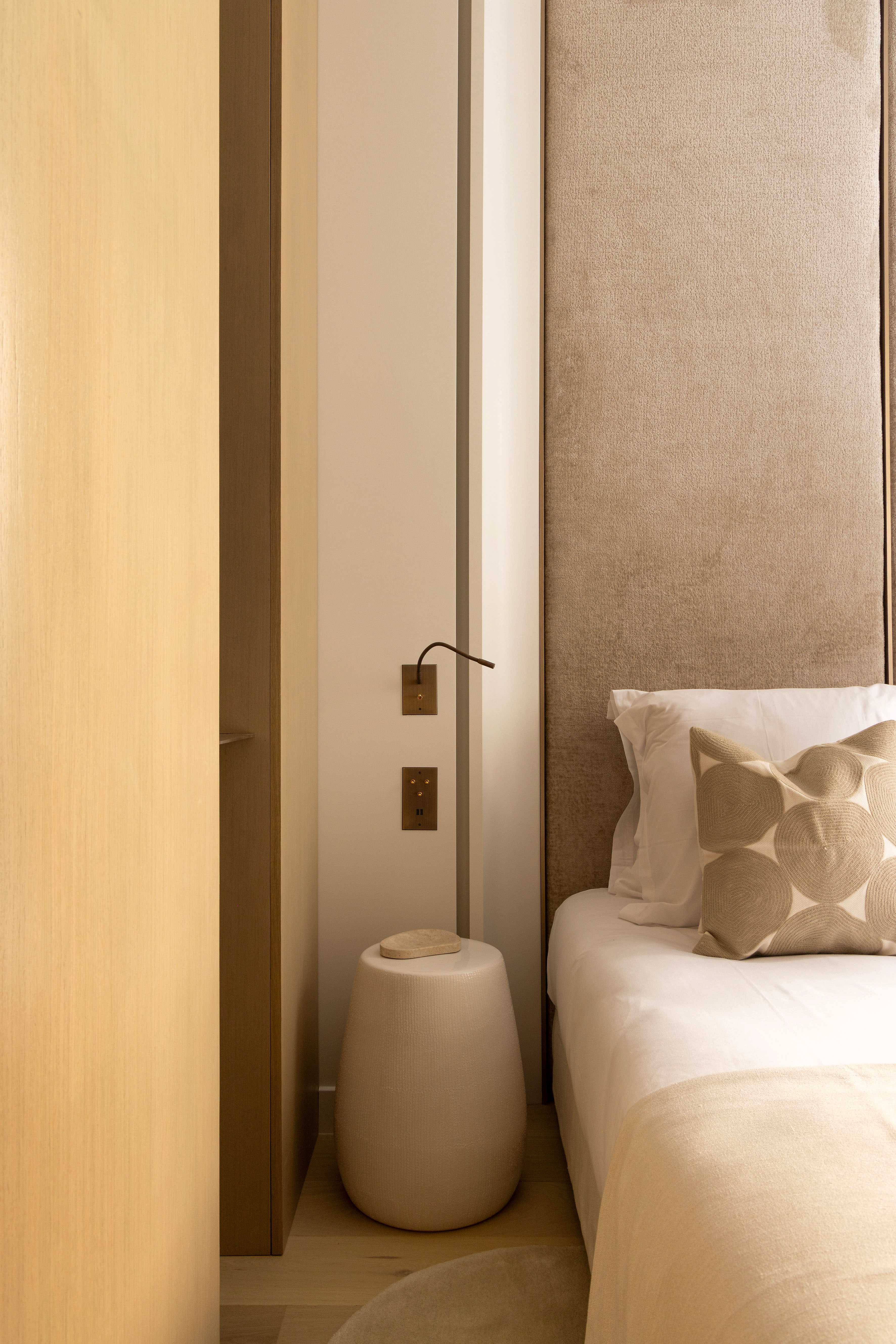 Détail du chevet avec table d'appoint en céramique beige, liseuse murale et tête de lit velours