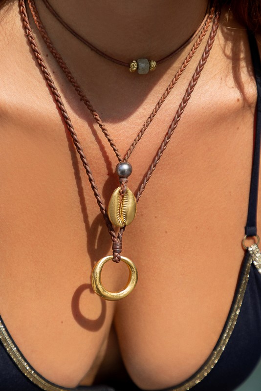 Island-inspired jewelry by Manos de Oro Saint Barth—featuring a handcrafted cowrie shell and gold ring pendant on braided leather, evoking soulful, sea-kissed elegance.