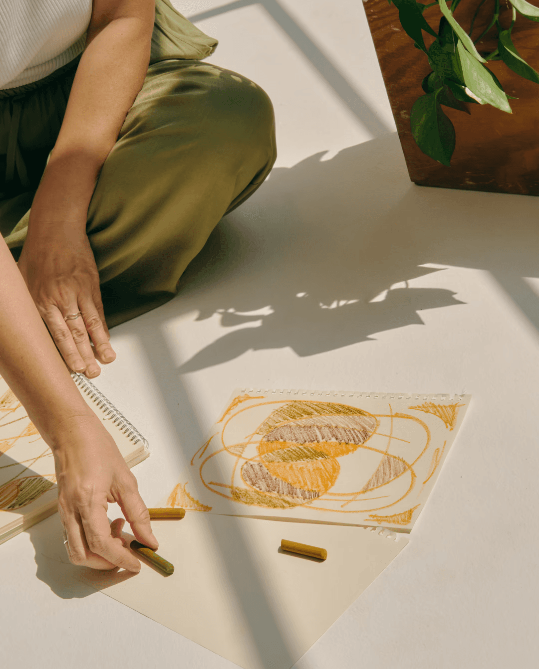 Girl sitting on the floor, drawing with orange and brown crayons on white paper, symbolizing the importance of mindfulness in mental and emotional wellbeing.