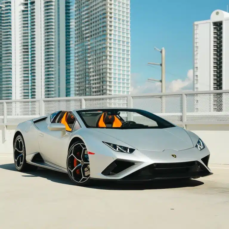 Front 3/4 view of a silver Lamborghini Huracán EVO Spyder with its convertible top down, parked with a modern city skyline in the background.