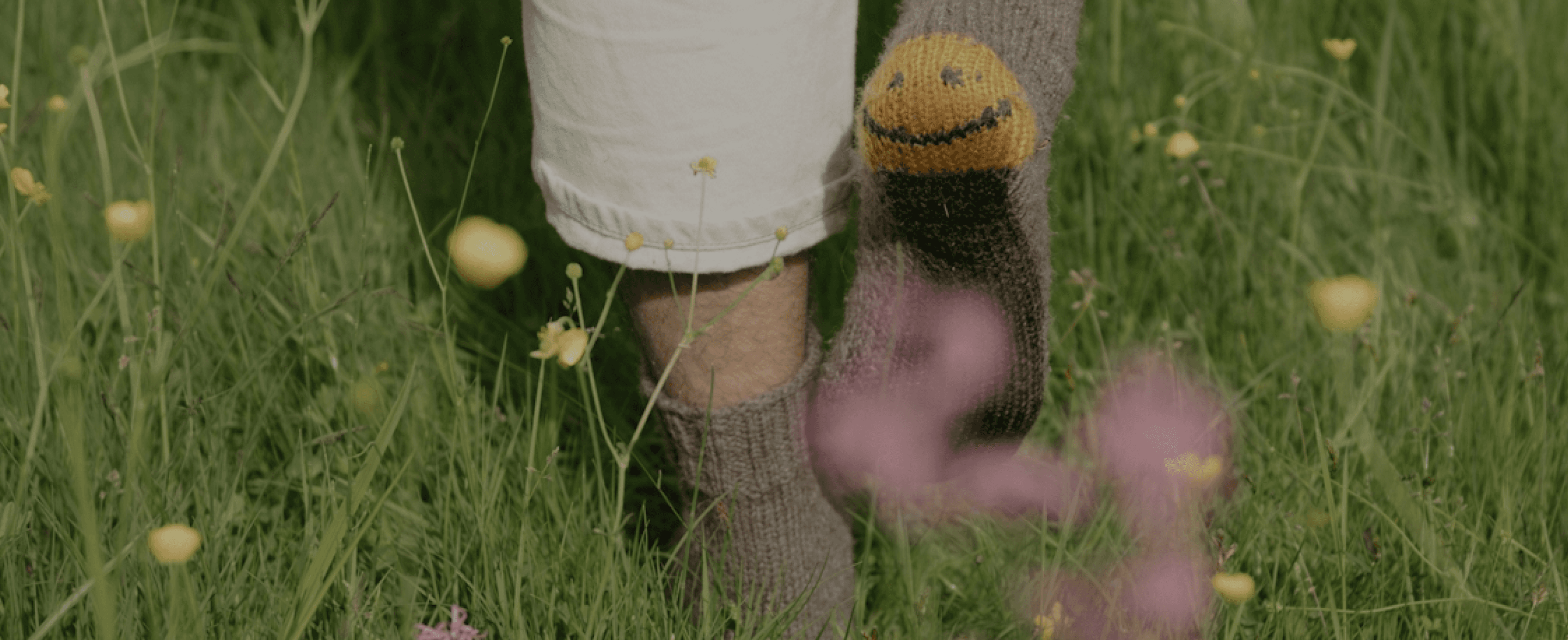 A person's lower body and feet standing in a grassy field filled with yellow and purple flowers, with a knitted sock or leg warmer featuring a smiley face design, creating a serene and whimsical visual composition.