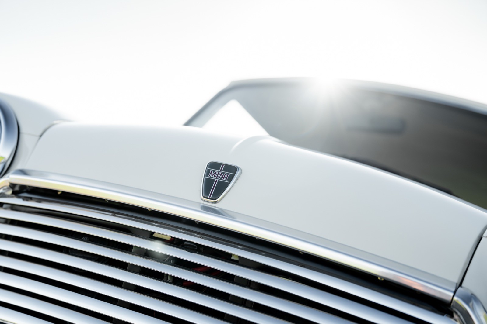 Close-up of a vintage car's grille and emblem, reflecting sunlight, set against a bright background.