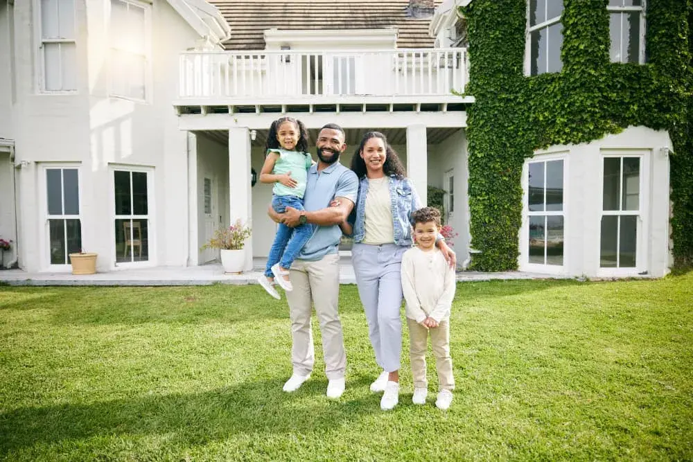A smiling family stands in front of a large white home covered in ivy, enjoying time together in the yard. The image represents stability, success, and the dream of homeownership made possible through Jumbo Loans from Chris Lewis Home Loans.
