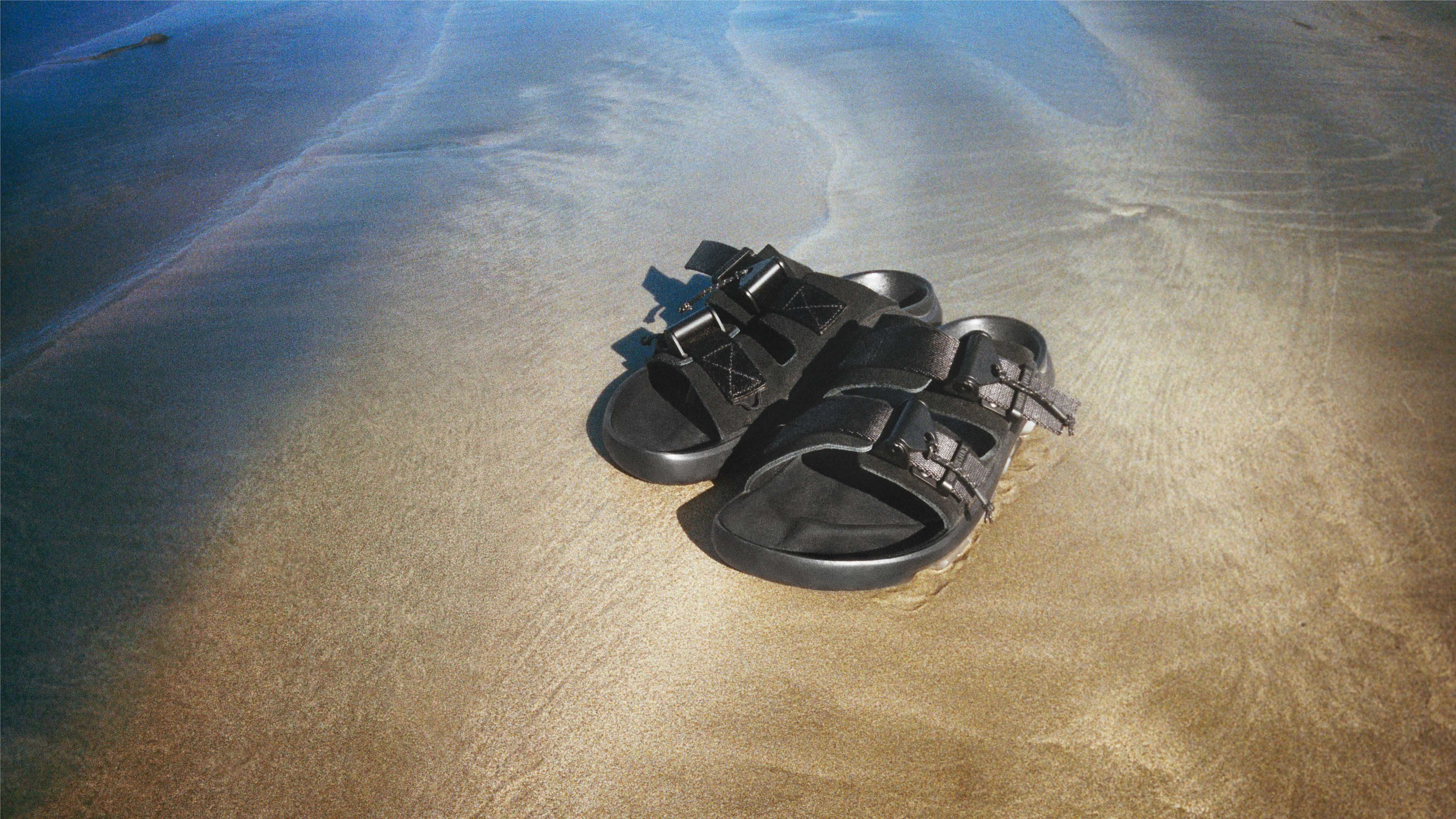 A pair of blackʹ Birkenstock-style sandals laid gently on damp beach sand, channeling the refined minimalism and heritage of the Birkenstock 1774 collection.