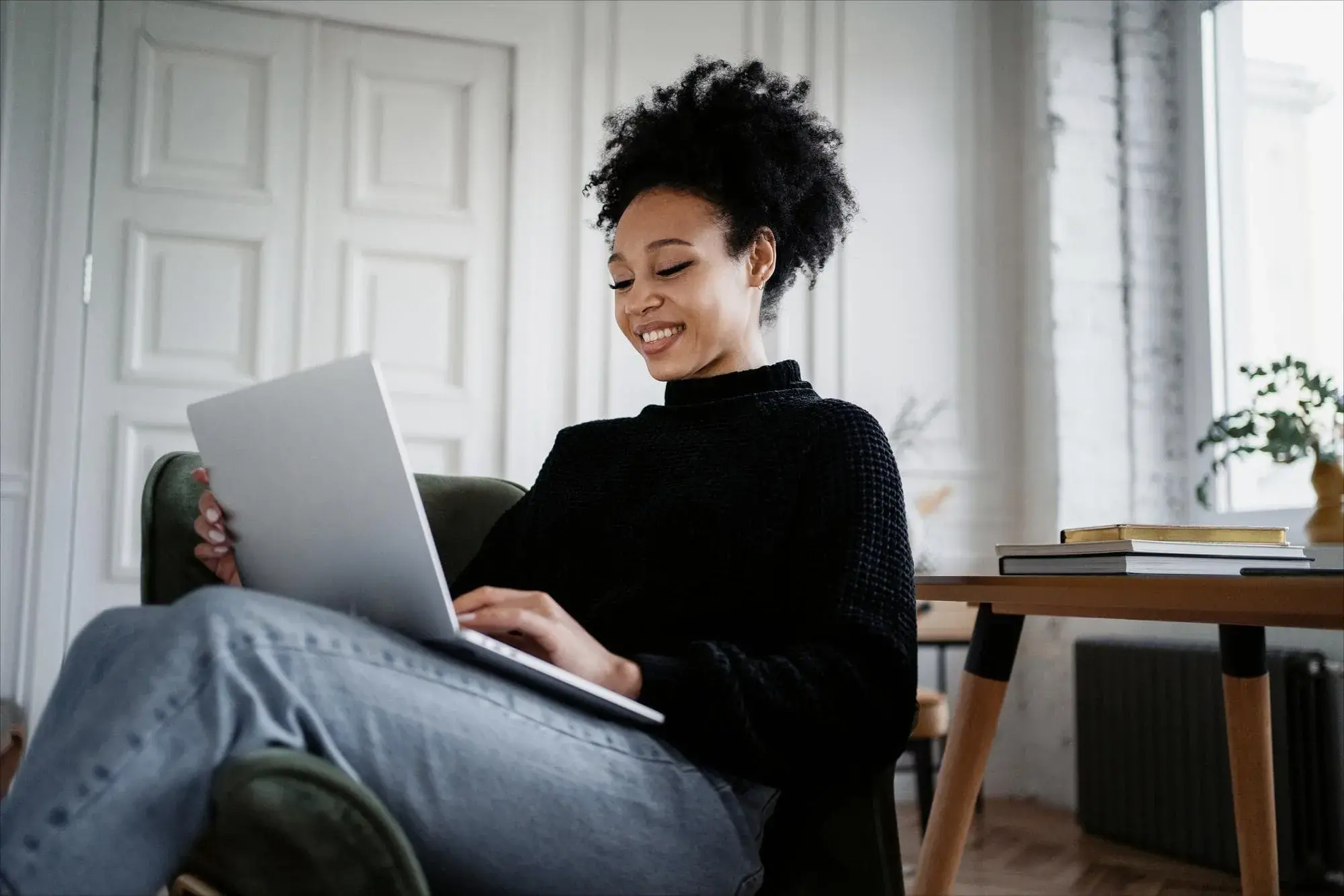 A woman sits comfortably at home using a laptop and smiling, surrounded by natural light and modern decor. The image conveys ease and confidence while exploring home loan options with Chris Lewis Home Loans.
