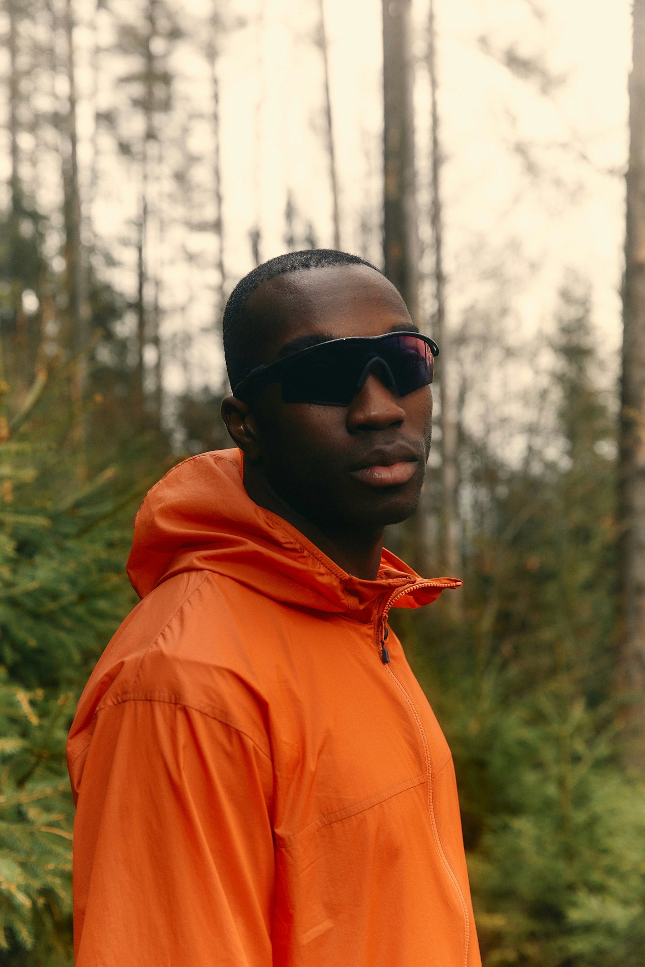 A man wearing an orange jacket and sunglasses standing in a forest.