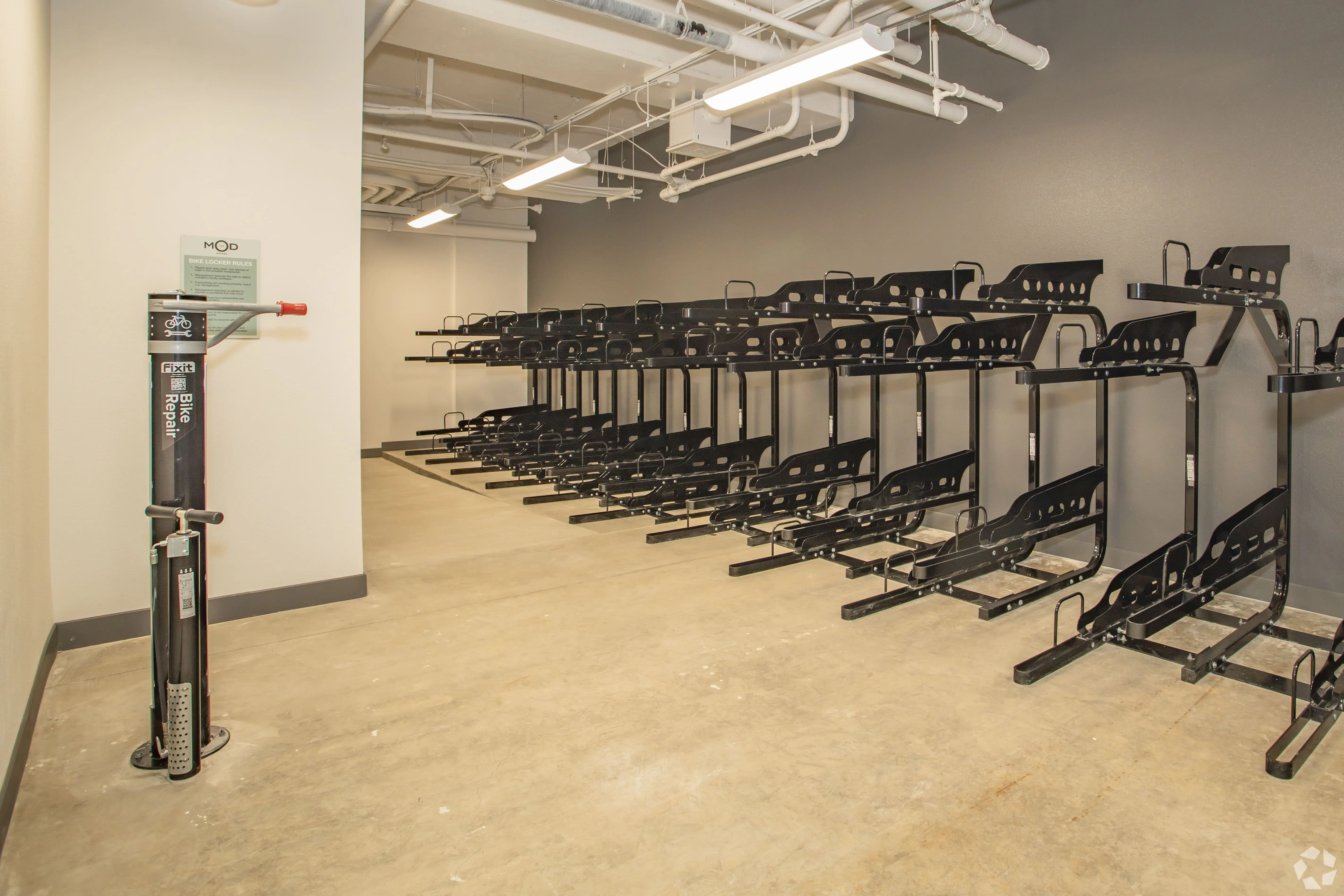 A clean, well-lit indoor bike storage room with multiple black wall-mounted bike racks on both sides epitomizes modern living spaces. Located in Midtown Sacramento, it features a bike repair station with tools in the foreground on the left. The floor is concrete, and the ceiling has exposed pipes and ducts.