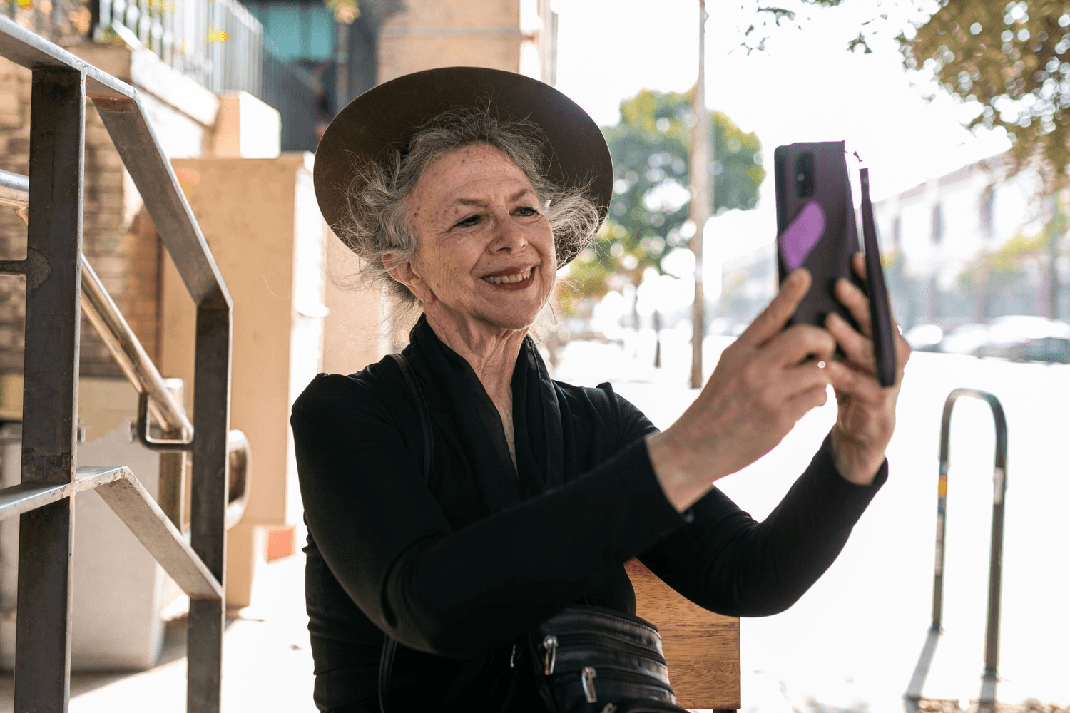 An older woman in a wide brimmed hat holding her phone up in front of her on a sunny street, smiling at the screen.