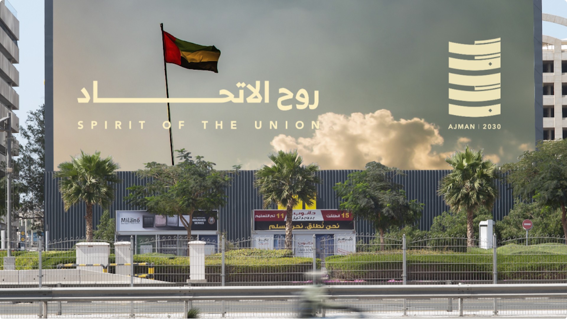 Billboard displaying a message, with a flag in the foreground and a cityscape in the background for Ajman2030