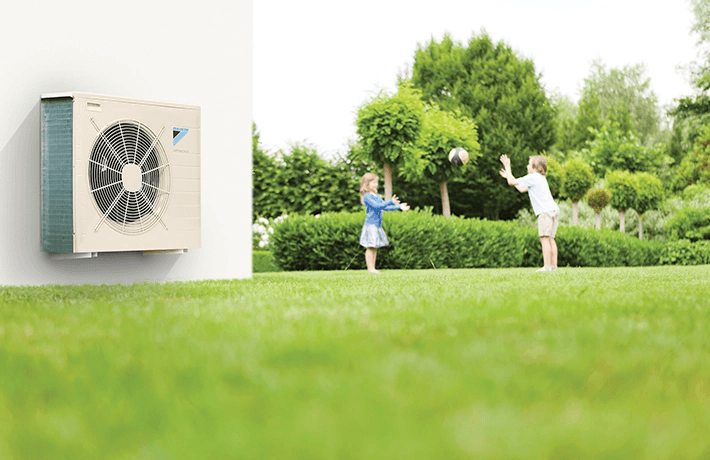 Vonkajšia jednotka klimatizácie Daikin na fasáde domu, v pozadí deti hrajúce sa na záhrade