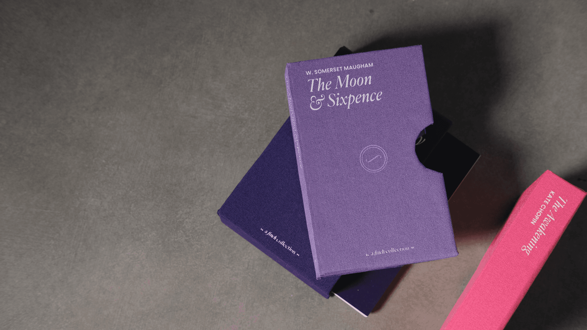 A purple book titled “The Moon & Sixpence” by W. Somerset Maugham rests on a dark gray surface. A dark blue book lies beneath it, and a pink book titled “The Awakening: Kate Chopin” is positioned to the right. A small circular logo is visible on the purple book’s cover.