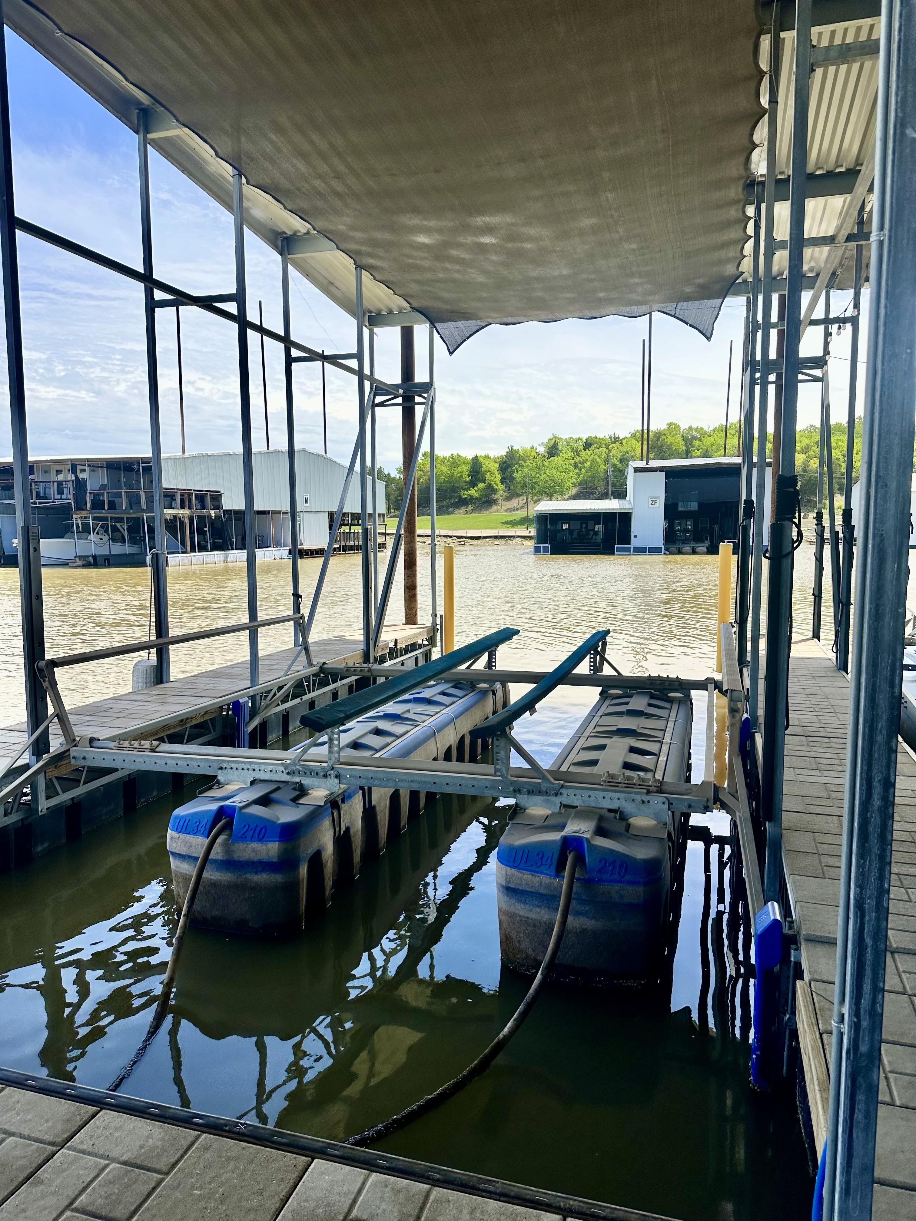 A boat dock with metal framework and floating platforms is situated on a calm lake, under a clear blue sky, with tree-covered hills and boathouses in the background.