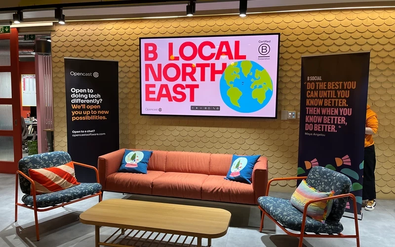 Modern seating area with a red sofa, two chairs, and a screen reading “B Local North East” with an Earth graphic.
