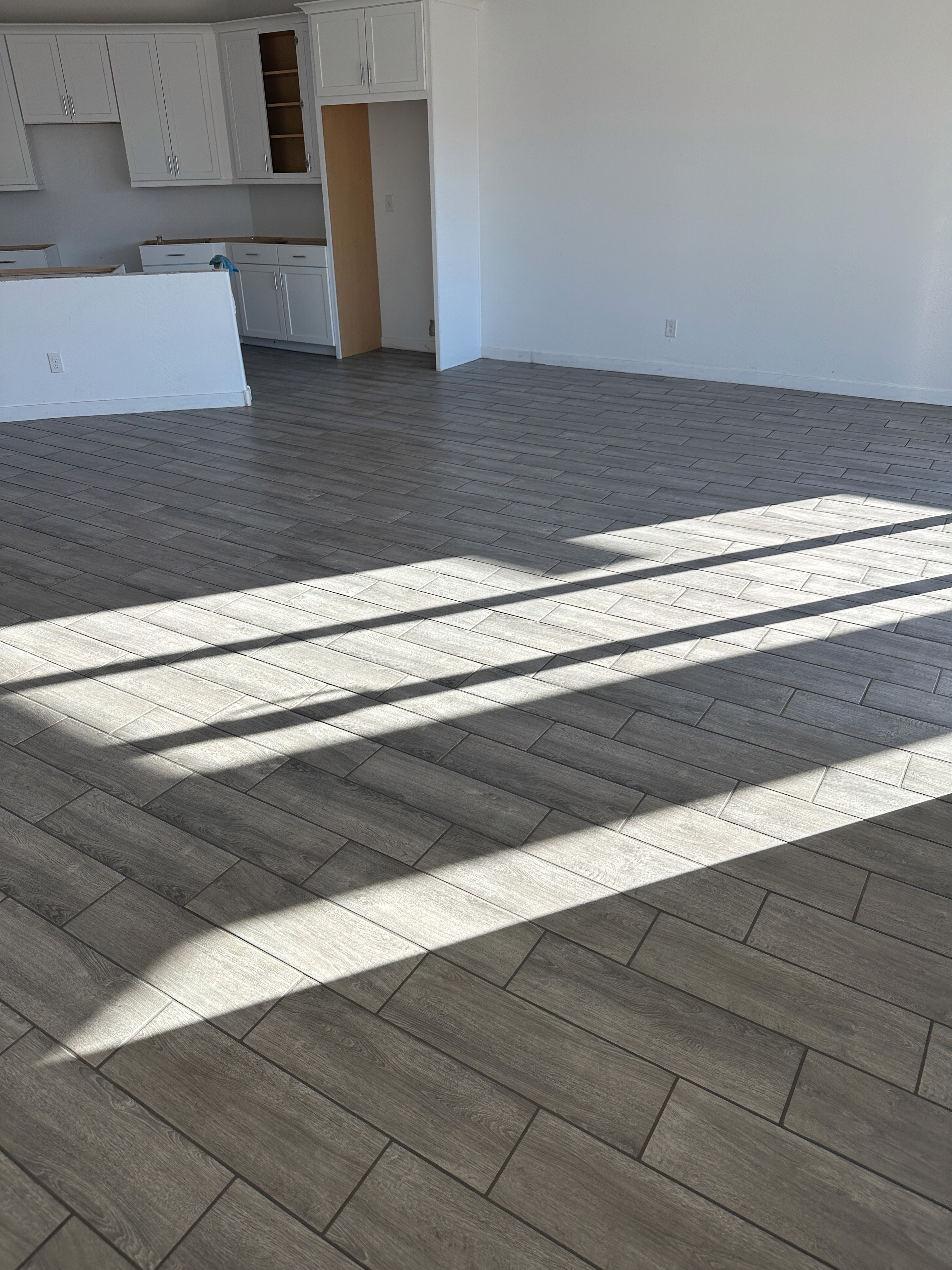 Modern kitchen and living area with wood-look tiles, large windows casting shadows, and a partially installed kitchen island.