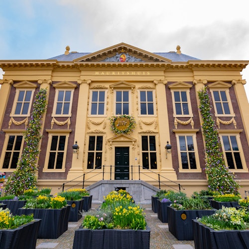 裝飾華麗的黃色和棕色建築，有三個入口、眾多窗戶，以及前面擺放著五顏六色的花卉的花槽。