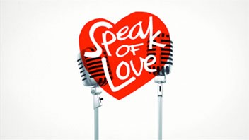 A red heart-shaped sign with the words "Speak of Love" in white cursive script is mounted on two silver poles against a plain white background. Two black-and-white microphones are positioned behind the sign, partially overlapping it.