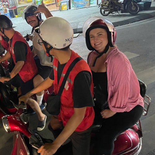 To personer med hjelme sidder på en scooter om natten; passageren, en smilende kvinde, har en lyserød skjorte på.