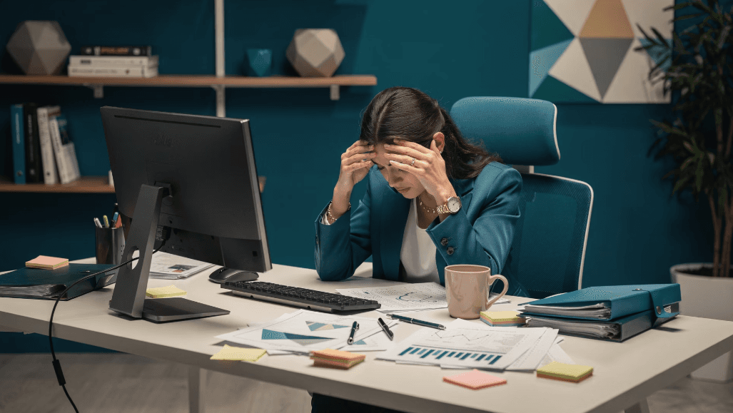 Frau sitzt gestresst am Schreibtisch und hält sich den Kopf zwischen Arbeitsunterlagen und Laptop