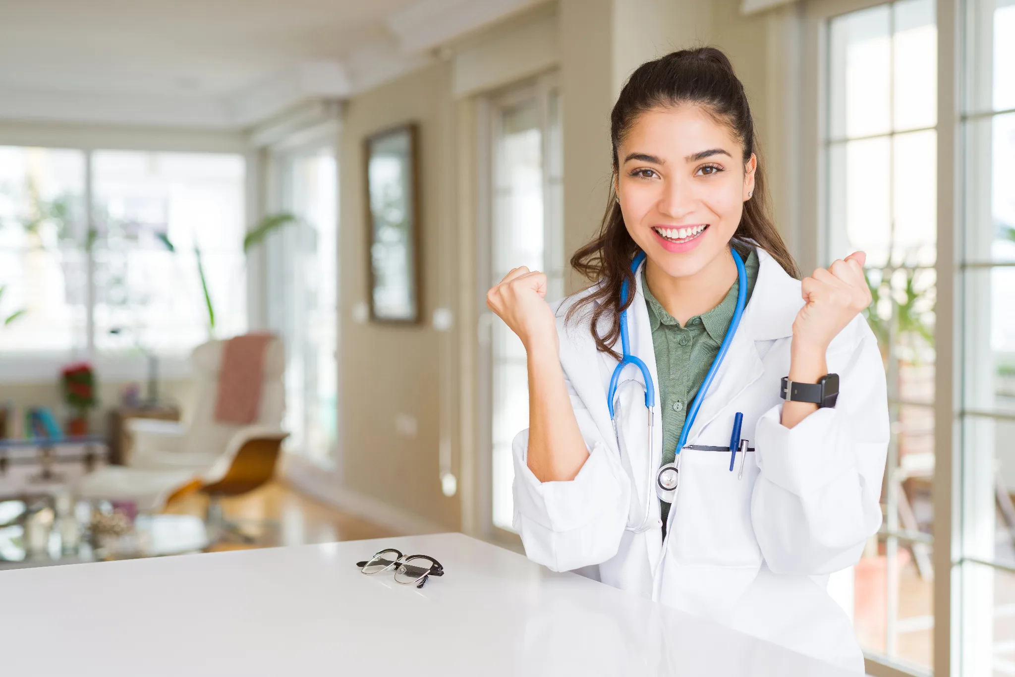 A young female doctor in a white coat and stethoscope smiles enthusiastically while giving a thumbs-up gesture at her bright, modern clinic office.