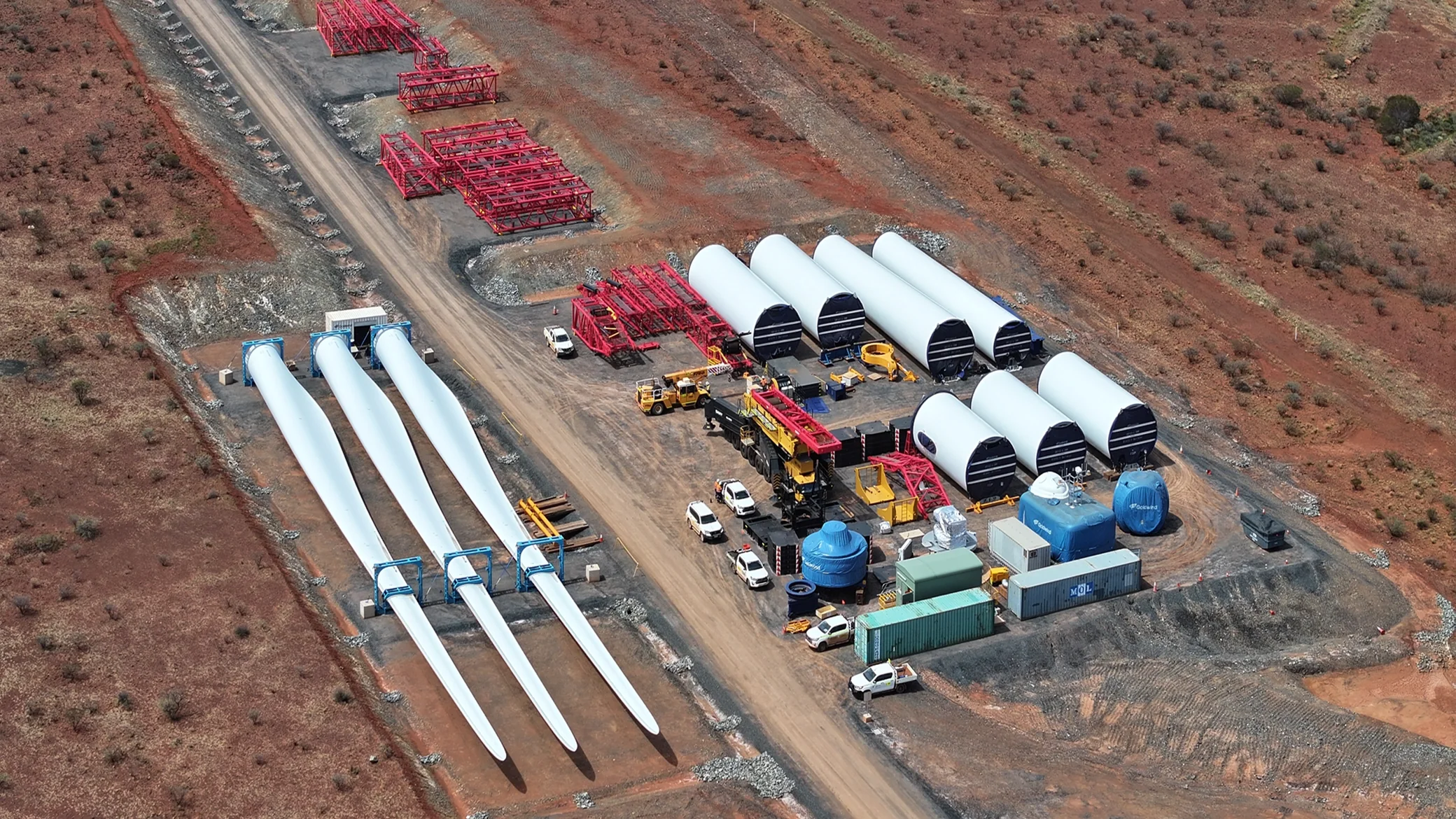 Aerial overhead shot of an industrial staging ground featuring wind turbine blades, tower sections, red structural steel framing, and heavy vehicles on red earth, highlighting sustainable net-zero energy infrastructure.