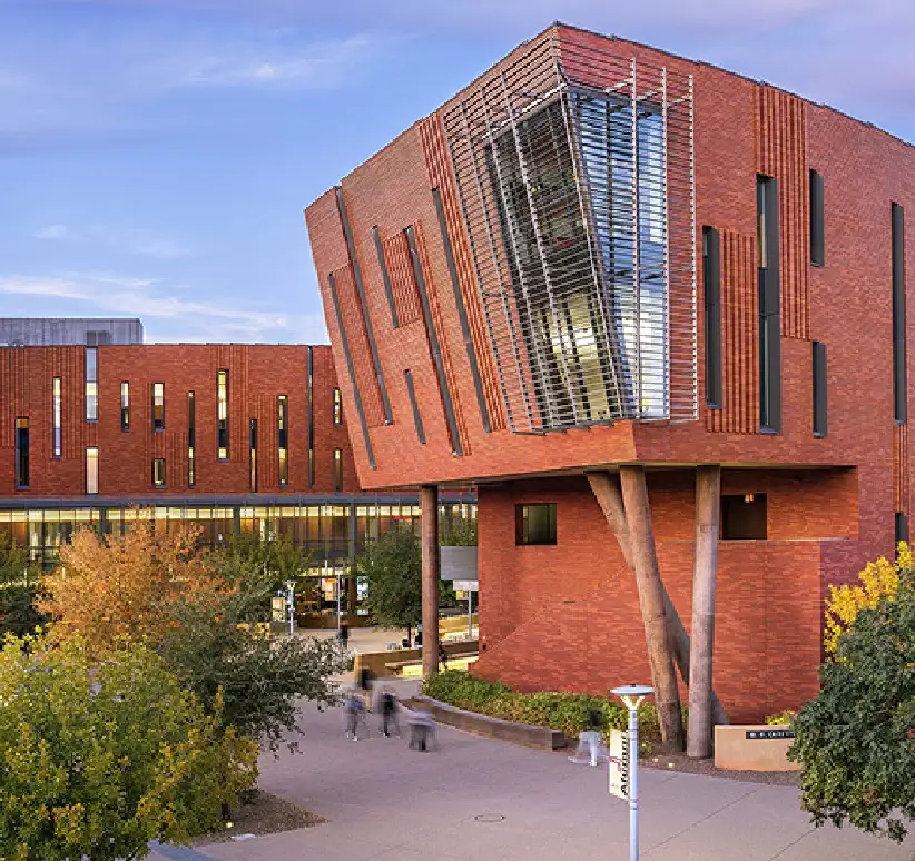 Edifício moderno de tijolos vermelhos com grandes janelas, cercado por vegetação e um caminho sob um céu claro.
