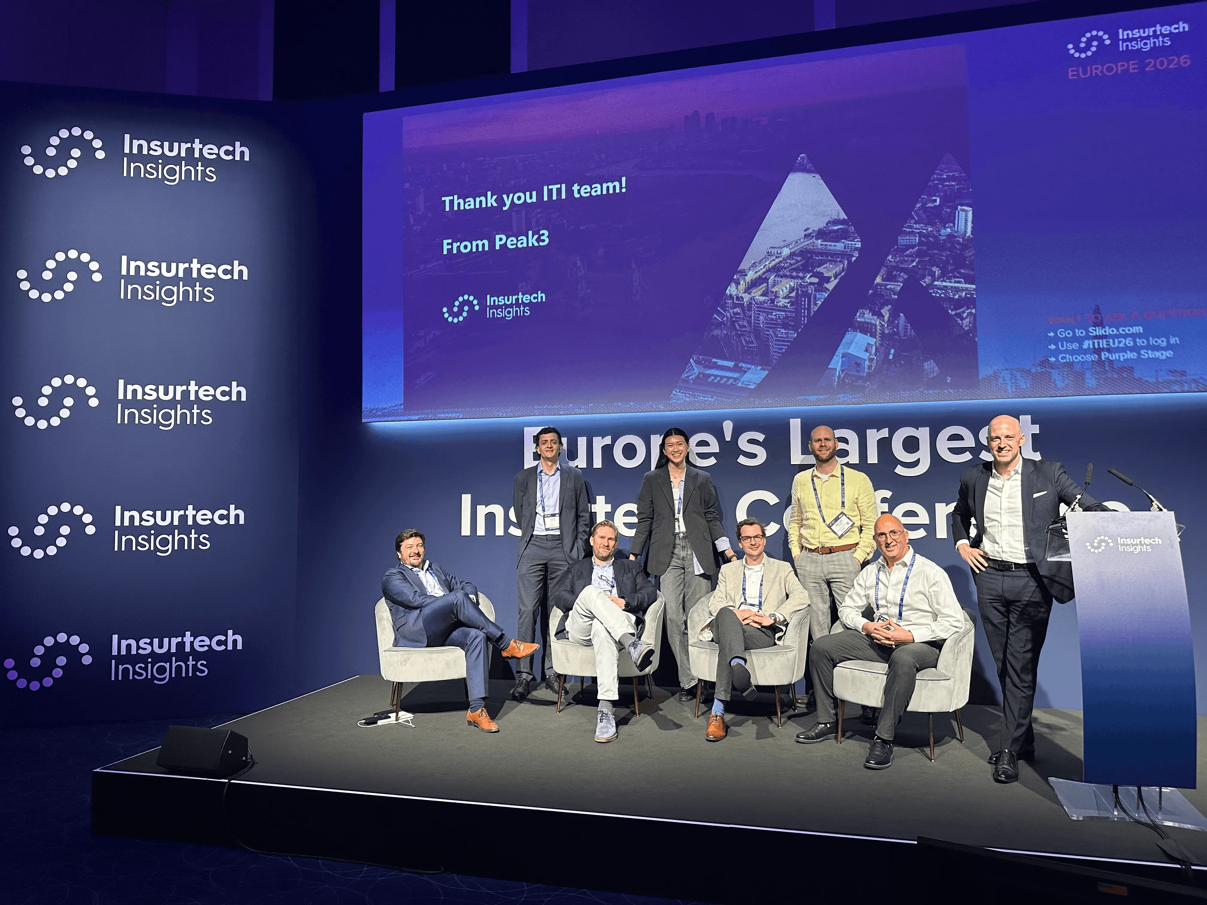 Panel discussion on stage with multiple speakers, featuring a blue backdrop and seating setup.