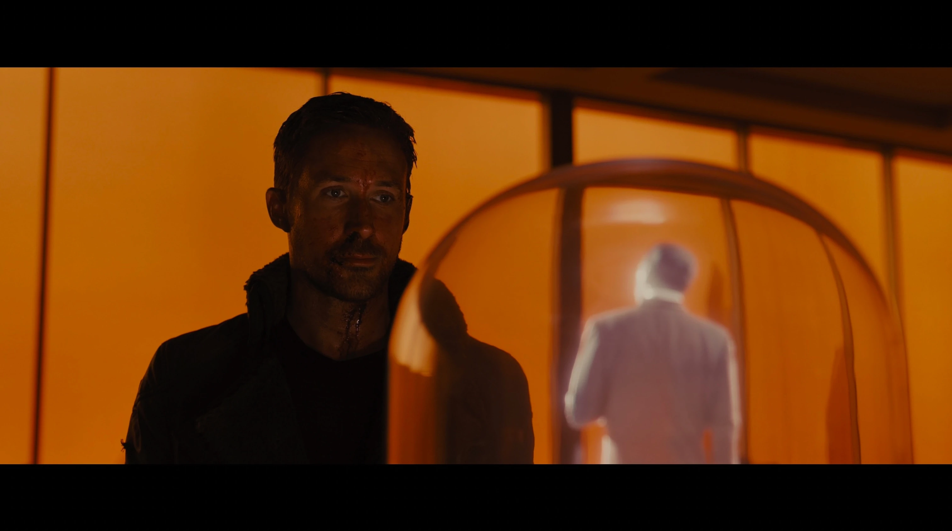 A man in dark clothing facing the camera beside a glowing white holographic figure seen from behind within an arched enclosure, bathed in deep orange light