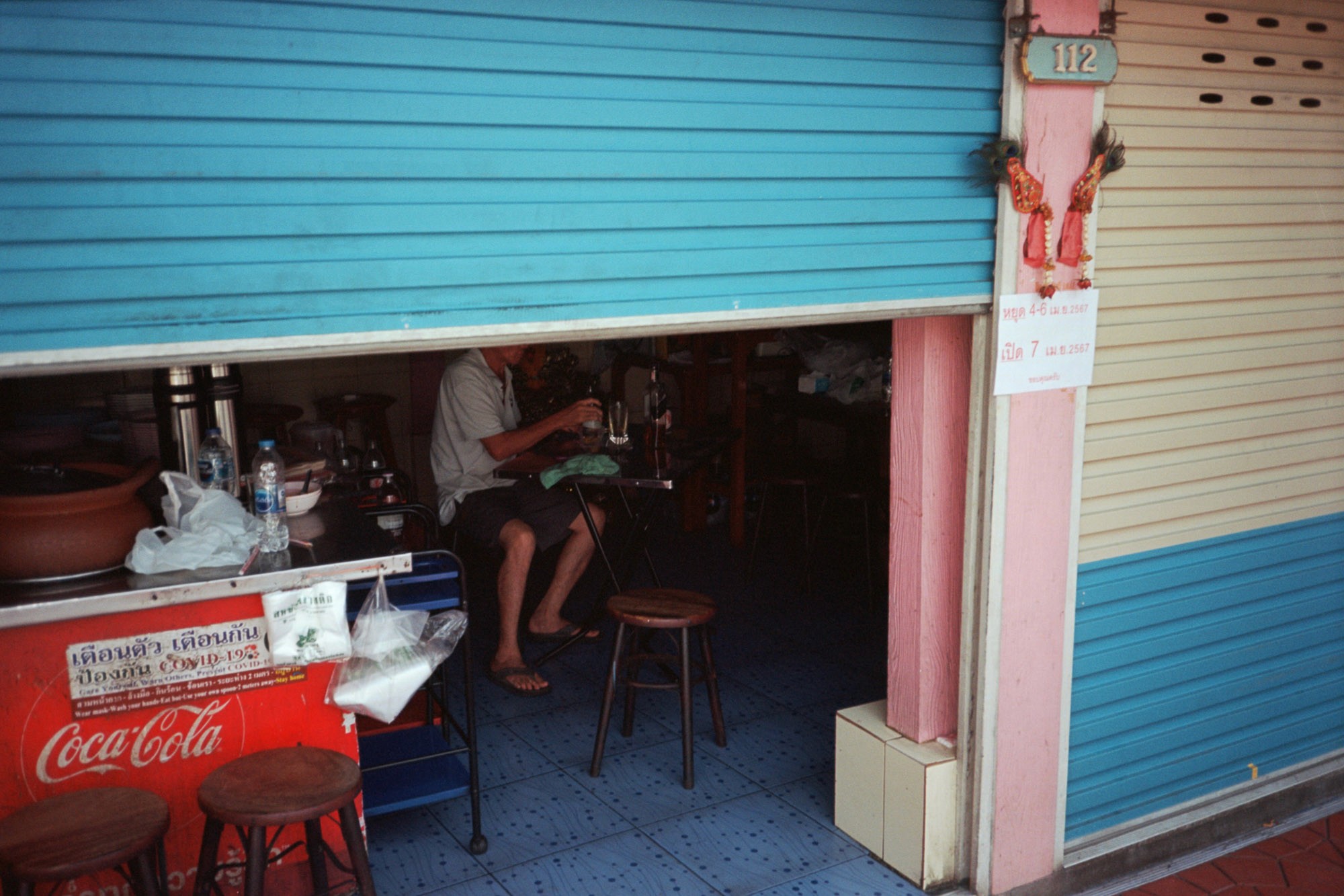 A person sits inside a small, partially open shop with blue tiled flooring, surrounded by a brightly colored storefront with blue and pink shutters, and a Coca-Cola branded red counter displaying bottled drinks, creating a vibrant street scene.