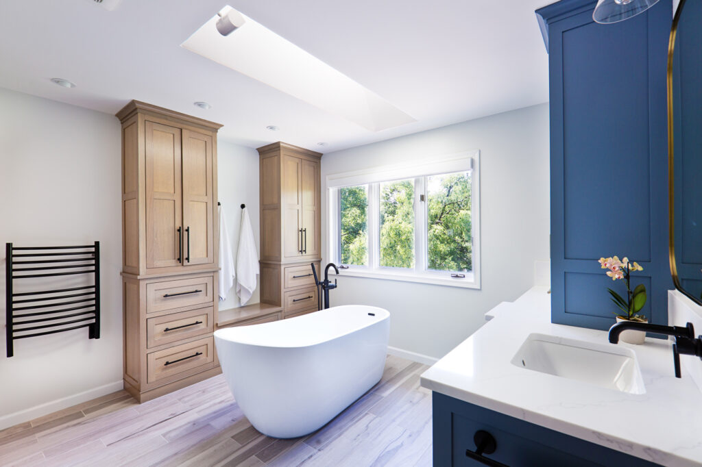 Bright modern bathroom featuring a white freestanding tub, light wood floors, large skylight, and built-in wooden and blue cabinetry. A walk-in glass shower with black fixtures sits near a private toilet room.