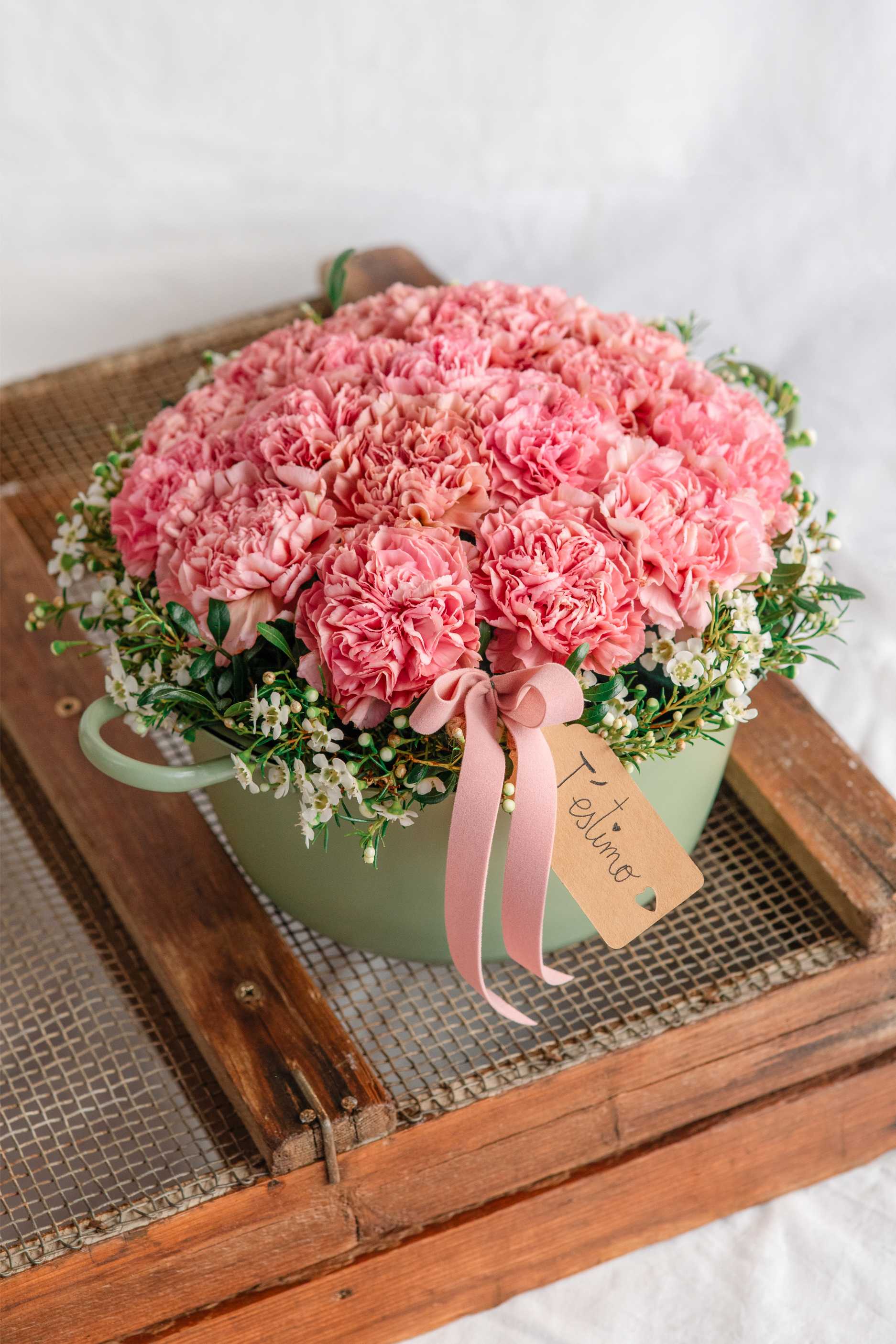 Centre floral amb clavells rosats disposats densament dins una base de metall verd suau, decorat amb cinta rosa i etiqueta amb el missatge “T’estimo” sobre una safata de fusta.