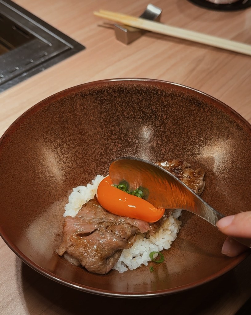 A bowl containing grilled beef, a raw egg yolk, and sliced green onions on a bed of white rice, with chopsticks and a grill in the background.