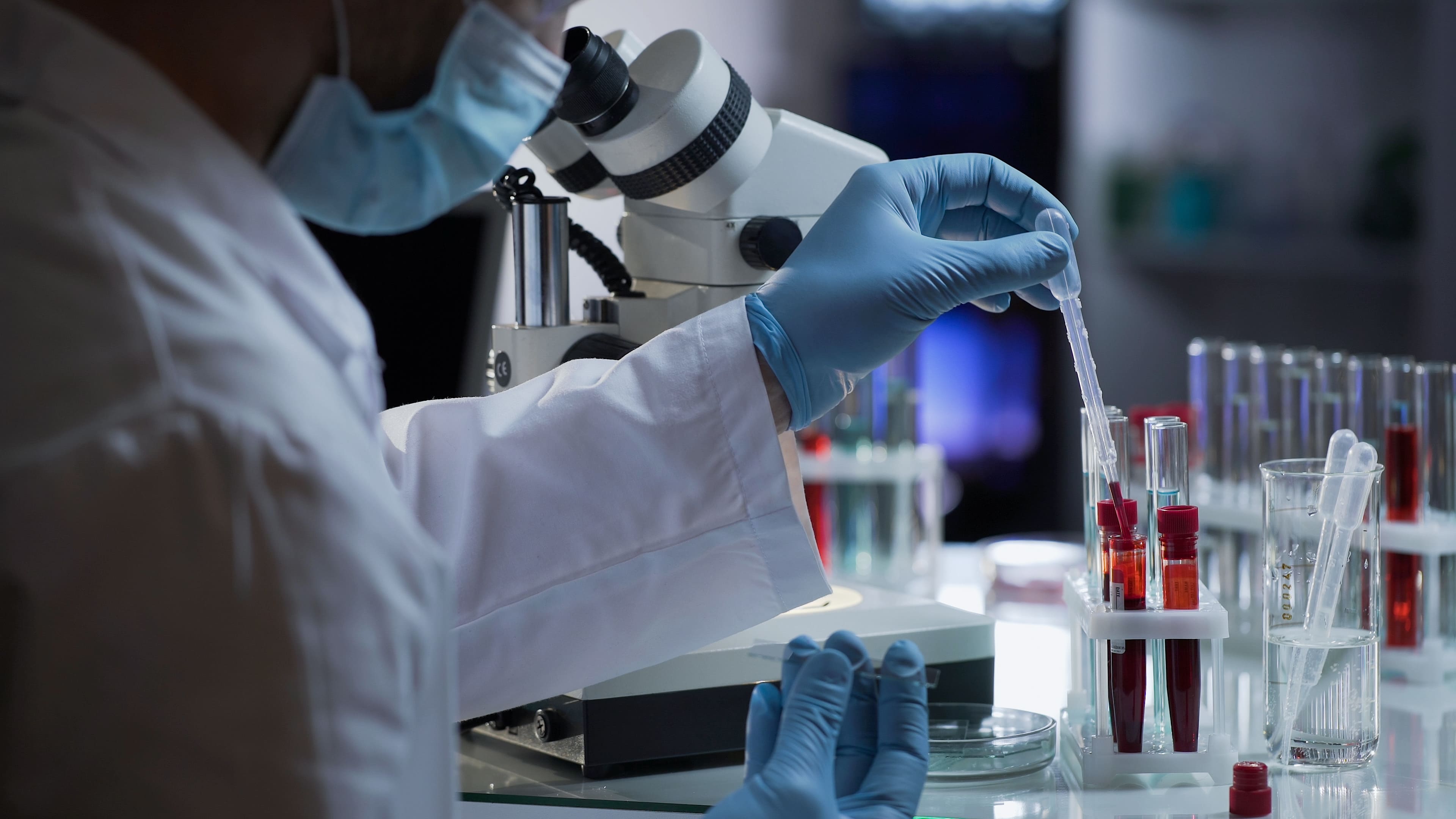A scientist in a laboratory carefully uses a pipette to transfer a red liquid into a test tube, with a microscope in the background, illustrating precision in scientific research and medical analysis.