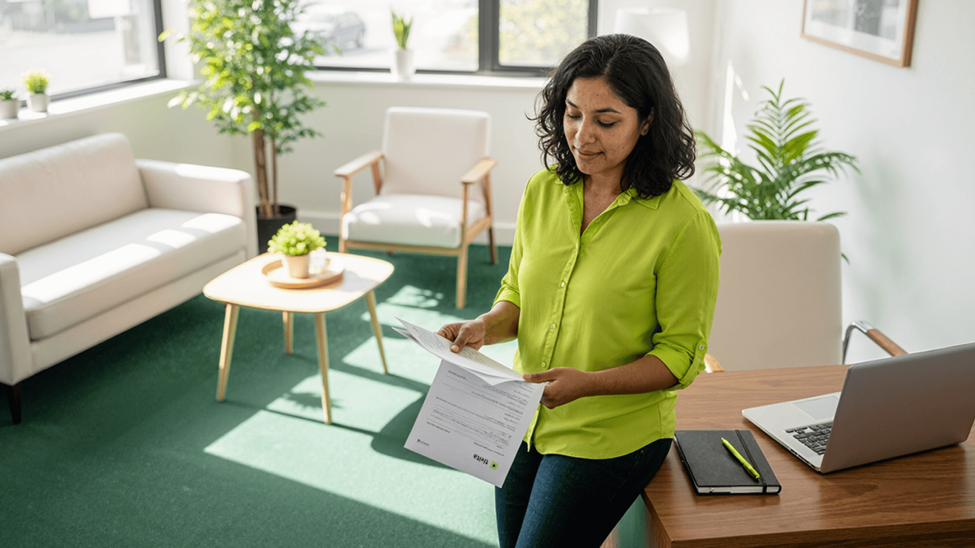 Profissional em ambiente de clínica segura e bem iluminada revisa documentos com o logotipo da Tivita sobre uma mesa de madeira, onde também estão um notebook e um caderno com caneta verde, representando o uso de prontuários e assinaturas digitais na gestão das clínicas.