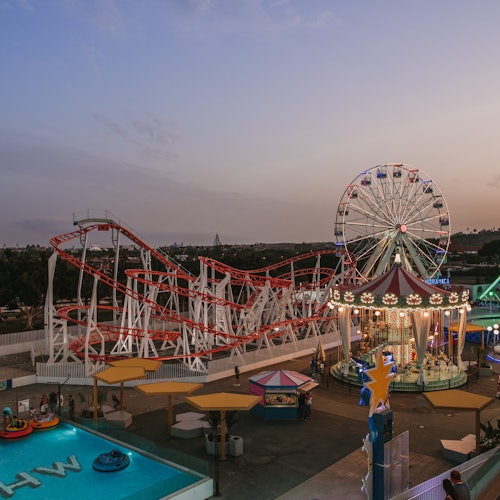 Een pretpark in de schemering met een achtbaan, reuzenrad, draaimolen, botsboten en kleurrijke lichten die het gebied verlichten.