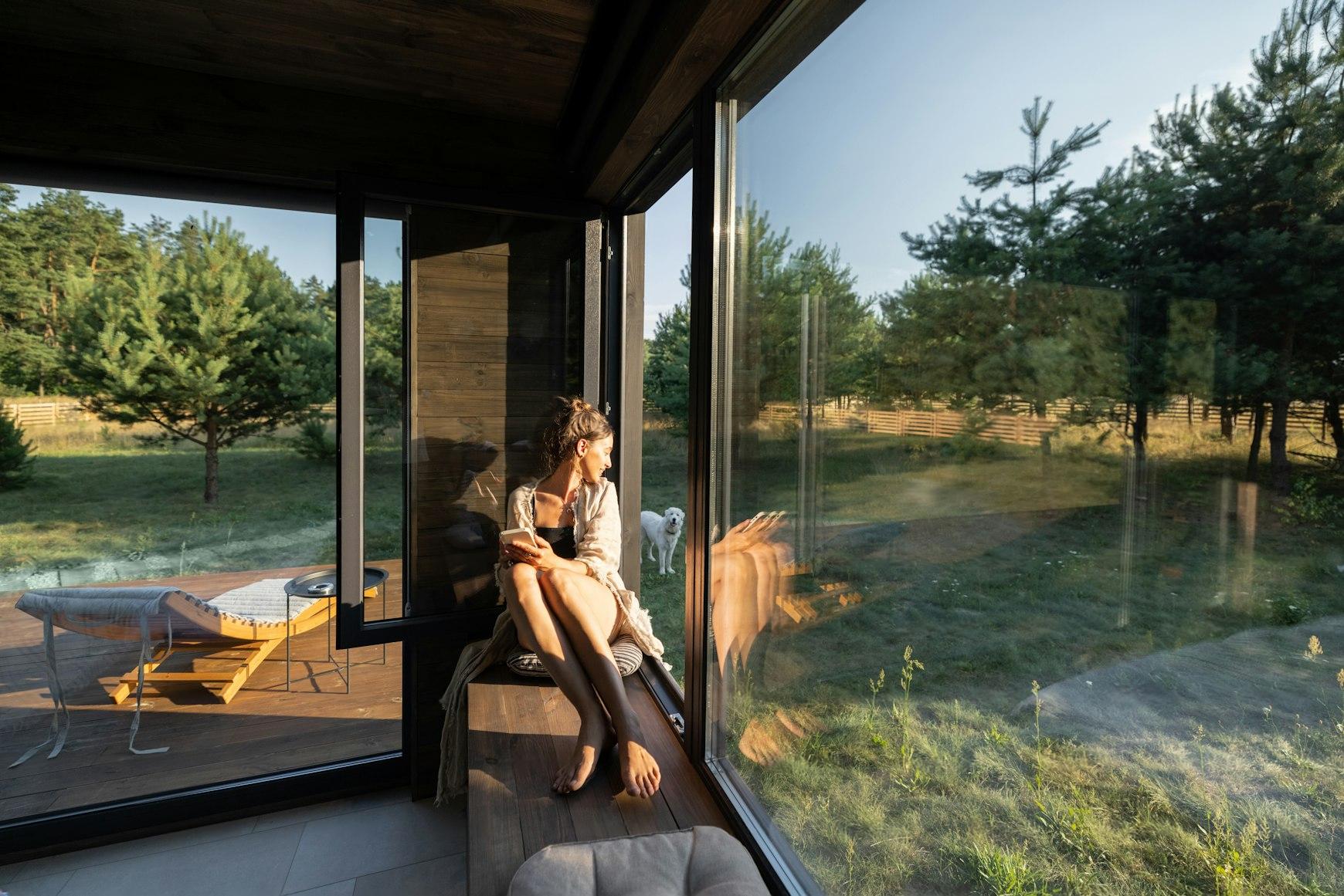 Woman reading on a window ledge inside a Strawtown Stack One modular home, with floor-to-ceiling glass panels overlooking a forested garden and a white dog outside