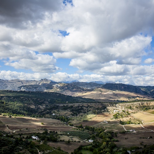 Laajat maaseutumaisemat peltoineen, hajallaan olevine puineen, pienine rakennuksineen ja etäällä näkyvine vuorineen puolipilvisen taivaan alla.
