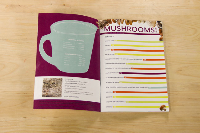 An open cookbook displays a page titled "Mushrooms!" with a list of contents including "We love," "Vintage," "Fresh," "Simple Pan-Roasted," "Deeply Roasted," "Grilled," "Club Mushroom Soup," "Mushroom Sauce," "How to Grow Mushrooms in a Tiny New York Apartment," "Wisconsin," "Second," and "Nic Farmers' Market." A light teal mug illustration is on the left page, and a photograph is visible on the left page's lower-left corner. The book rests on a light wooden surface.