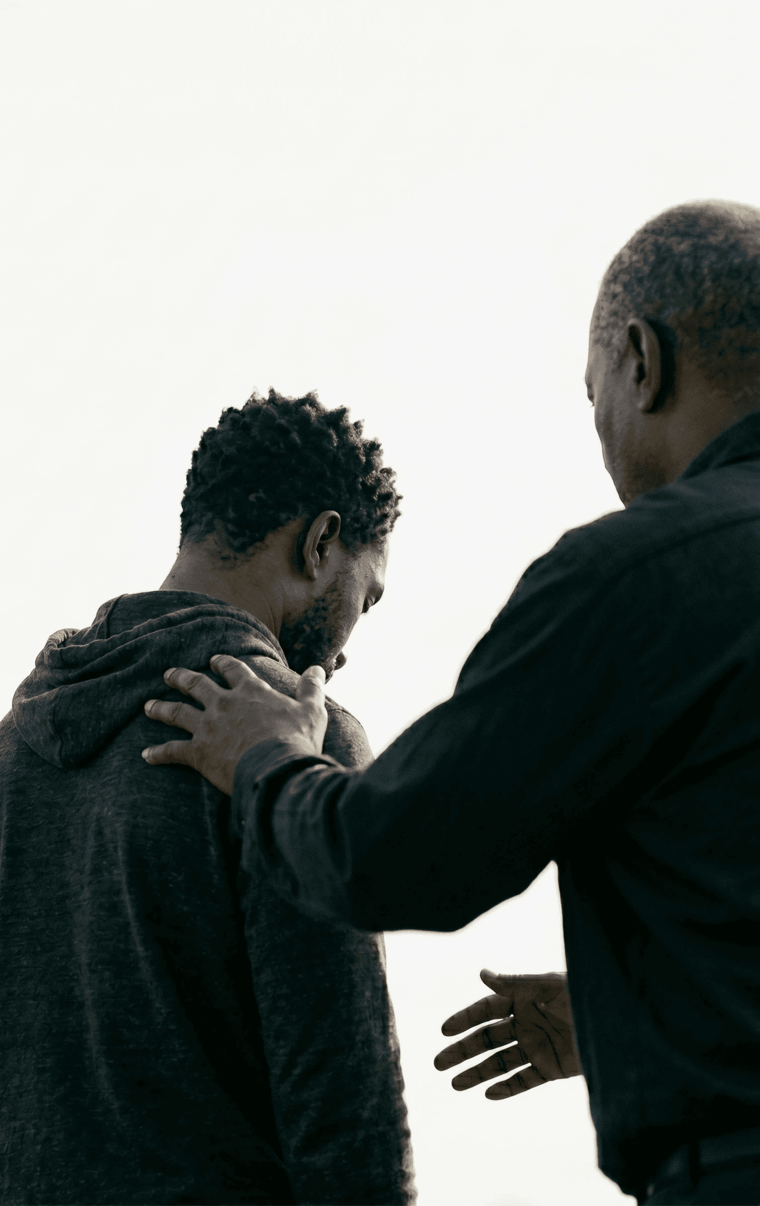 Two people are interacting outdoors; one person is gently placing a hand on the other's shoulder, conveying support and empathy on a clear day.