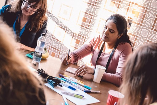 Group of people sitting around a table during a workshop session, with one person writing on a sticky note and various pens, markers and paper spread across the table.