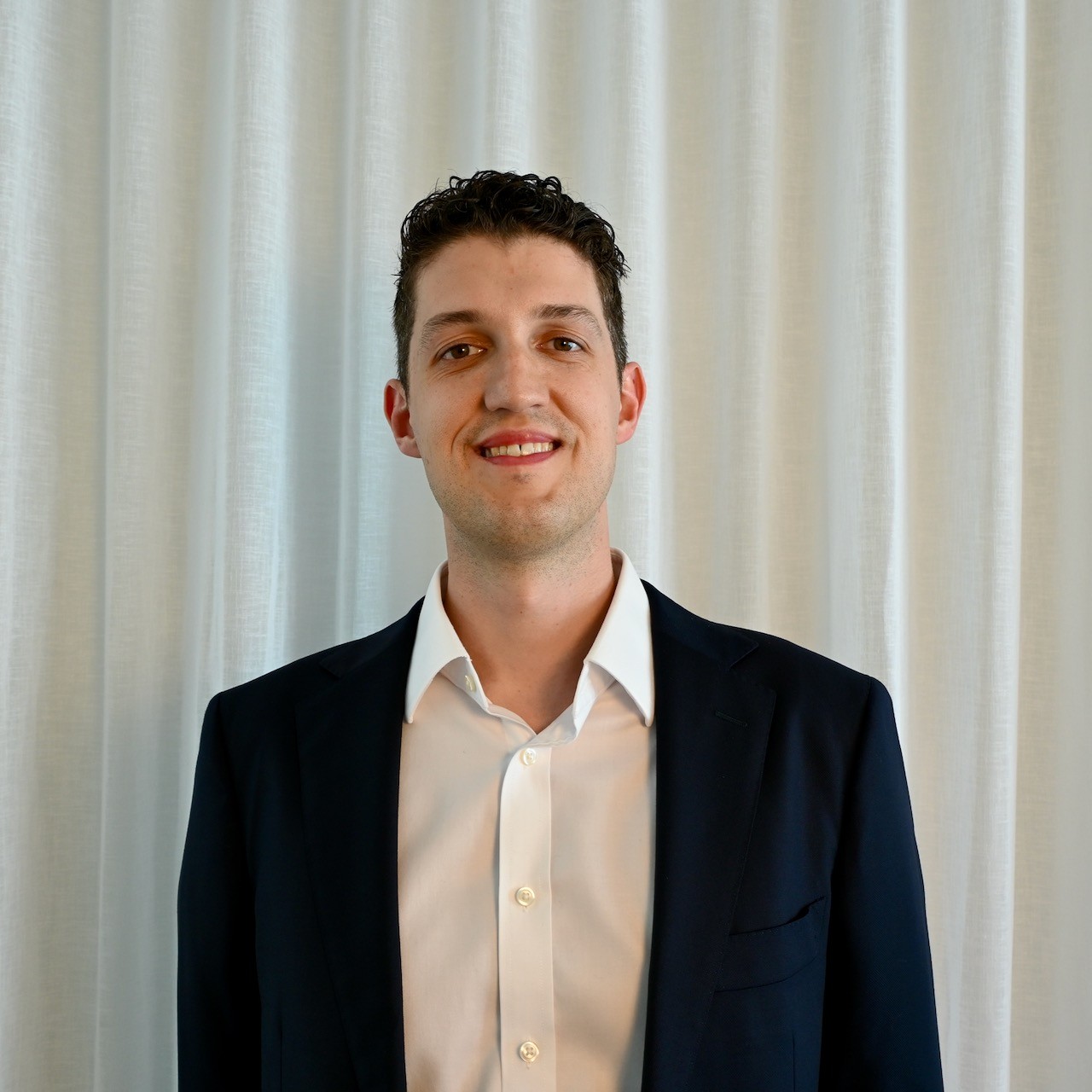 A smiling man with curly hair is sitting at a desk, looking at the camera, with a laptop and a lamp in the background.